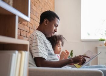 Side view of young Black father reading a book with his little daughter while sitting on the couch