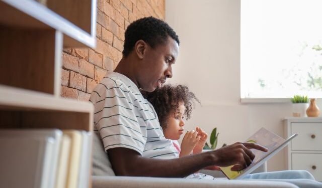 Side view of young Black father reading a book with his little daughter while sitting on the couch